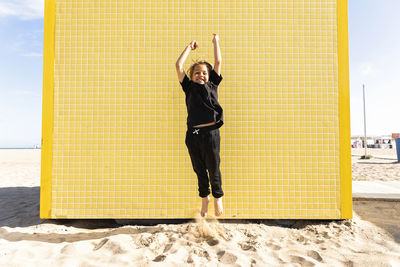 Cheerful boy jumping in front of yellow wall at beach