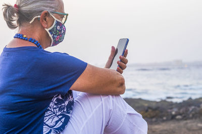 Side view of senior woman wearing mask using smart phone siting by sea