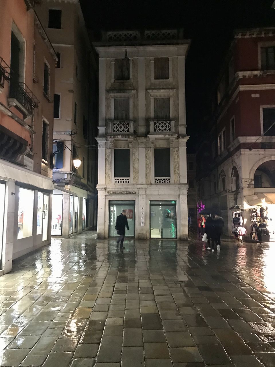 PEOPLE WALKING ON WET STREET AMIDST BUILDINGS