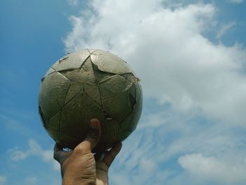 Cropped hand holding old soccer ball against sky