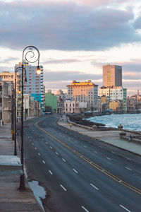 Road by buildings in city against sky during sunset