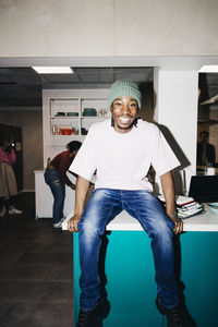 Portrait of happy young man sitting on dining table in kitchen at college dorm