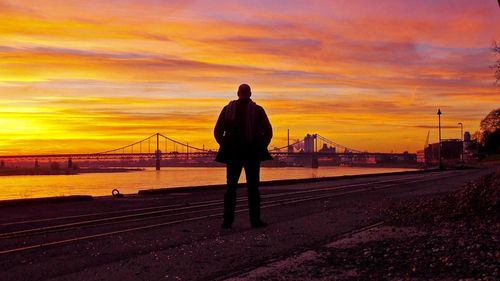 Rear view of silhouette man standing on bridge against sunset sky