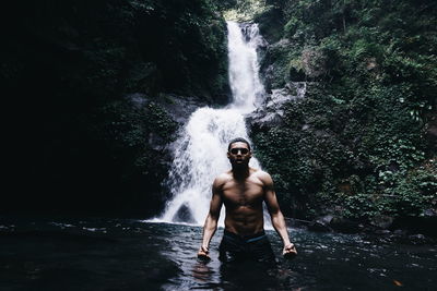 Portrait of shirtless man standing against waterfall