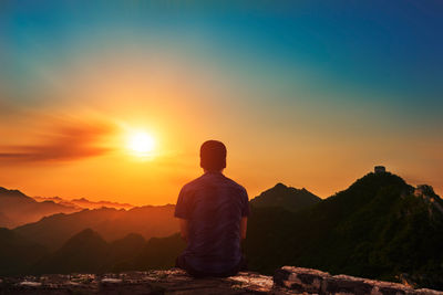 Rear view of man sitting on mountain against sky during sunset