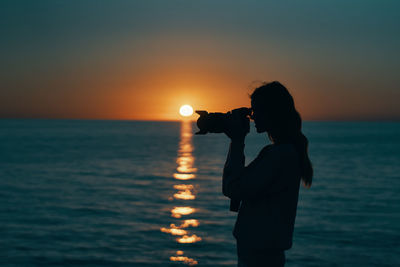 Silhouette man photographing sea against sky during sunset
