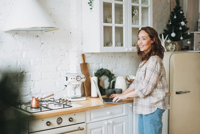 Attractive smiling woman with curly hair in plaid shirt with laptop near window at bright kitchen