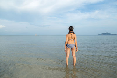 Rear view of girl in bikini standing at beach against sky