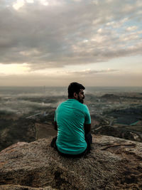 Rear view of man sitting on rock looking at sea against sky