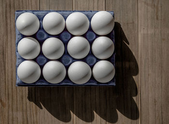 A dozen white eggs in a blue box on a wooden table. shadows.