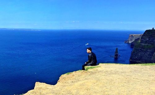Man sitting on rock by sea against clear blue sky