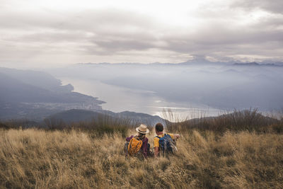 Happy mature couple sitting on grass in front of mountains under cloudy sky