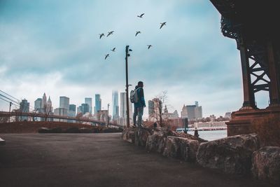 Full length of man standing on rock by river in city against cloudy sky