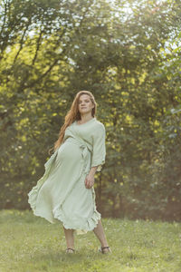 Dancing pretty regnant woman with big belly, green trees, meadow on background. white female wears