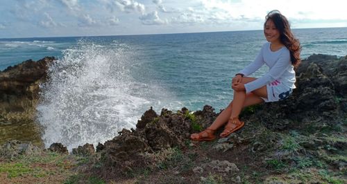 Full length of smiling woman sitting on rock at beach against sky