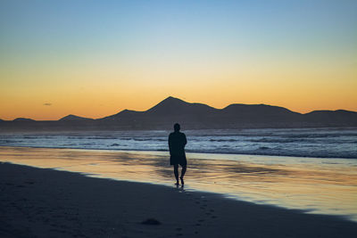 Rear view of man walking at beach against sky during sunset