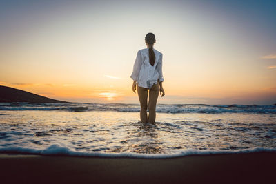 Rear view of seductive woman standing on shore at beach during sunset