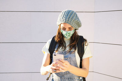 Young woman using phone while standing against wall