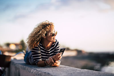 Young woman looking away while sitting on mobile phone against sky