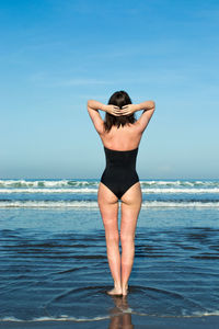 Rear view of woman in one piece swimsuit standing on shore at beach