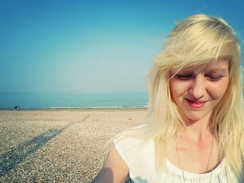 Close-up of blond woman smiling while standing at beach against blue sky