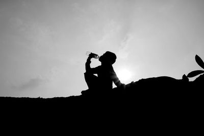 Side view of silhouette boy drinking water from bottle while sitting on land against sky