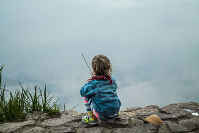 Rear view of girl sitting on rock against cloudy sky