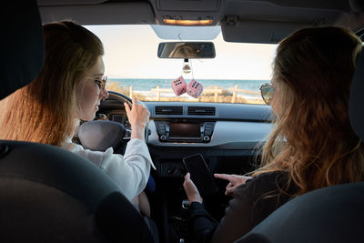 Back view of traveling women sitting in modern automobile and checking route on smartphone while navigating on online map during road trip