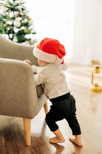 A little boy in a santa claus hat is standing near an armchair with a christmas tree toy i