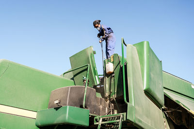 Low angle of unrecognizable employee in workwear and protective respirator cleaning agricultural machine in countryside on sunny day