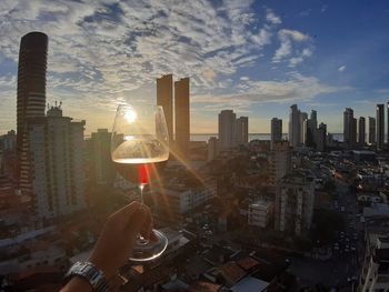 Man holding glass of buildings against sky during sunset