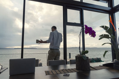 Rear view of man standing by window against sky