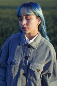 Upset pensive young female with blue hair dressed in trendy jacket standing in green field in sunny evening