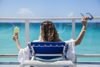 Rear view of woman having drink while relaxing on lounge chair against sea