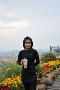 Portrait of smiling young woman holding coffee while standing on grassy field against sky