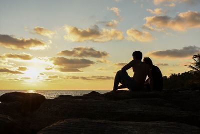 Men sitting on rock by sea against sky during sunset