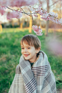 Portrait of a funny little boy wrapped in a blanket enjoying cherry blossoms in a city park.