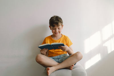 A cute boy wearing an orange t-shirt is sitting on a soft ottoman reading a book