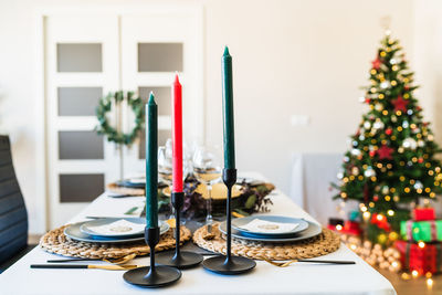 Festive table setting with candles located near fancy christmas tree with shiny garlands in spacious living room before family gathering