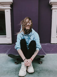 Full length of woman sitting on skateboard against wall