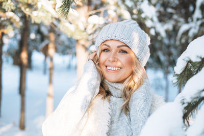 Smiling blonde woman with blonde hair in winter clothes hands near face in snowy winter forest