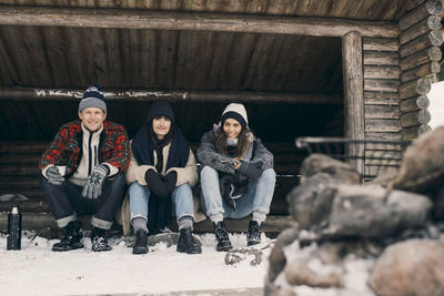 Full length portrait of friends sitting in log cabin against trees during winter