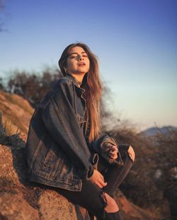 Side view portrait of young woman wearing denim jacket while sitting on mountain during sunset