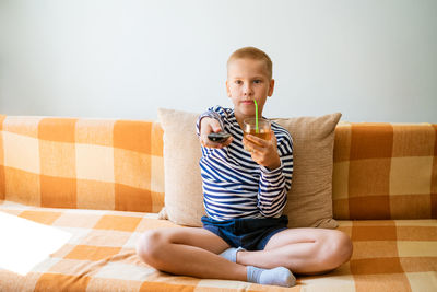 Caucasian school-age boy sits on a sofa in casual clothes with a remote control