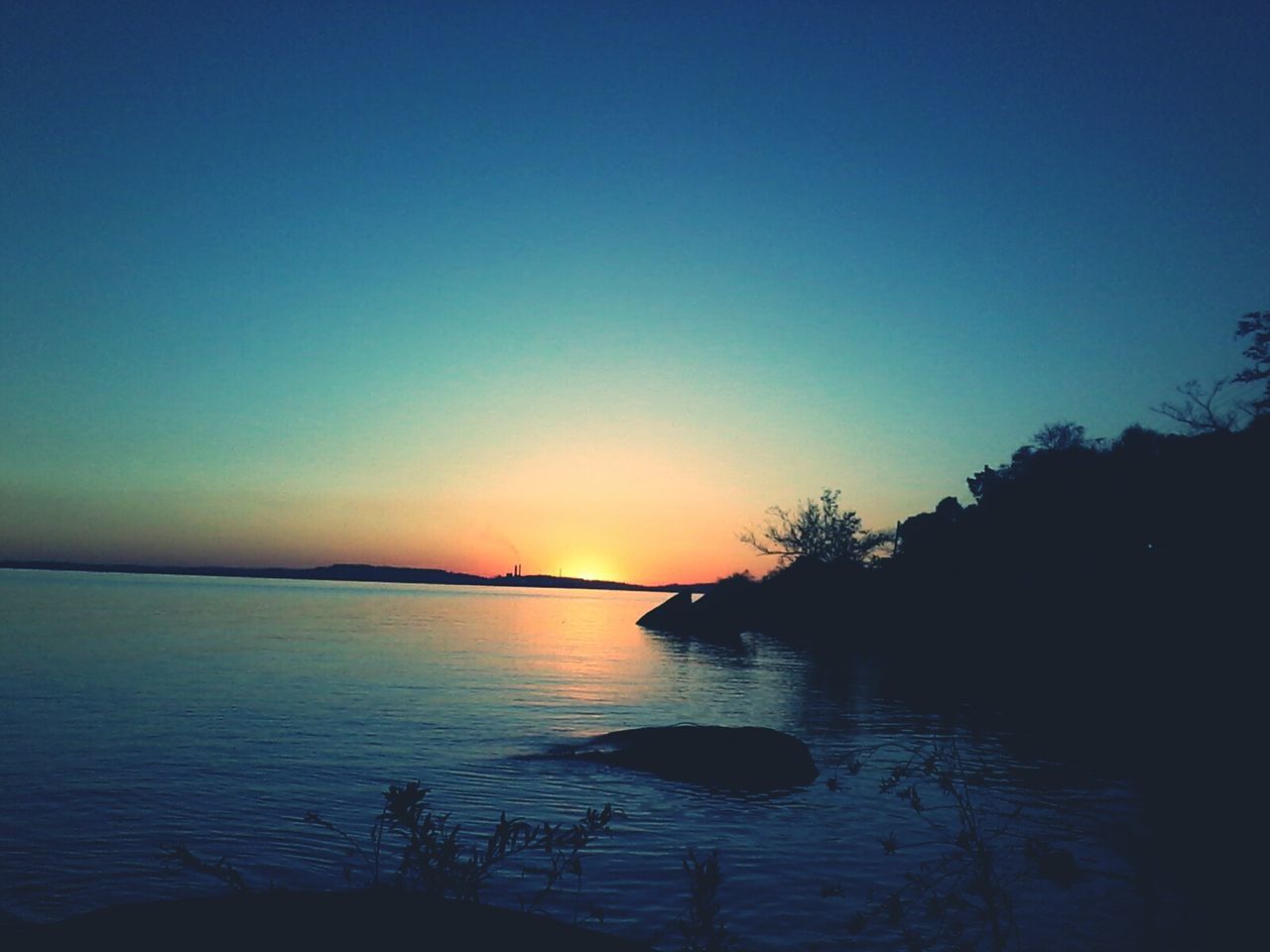 sunset, water, silhouette, tranquil scene, tranquility, scenics, beauty in nature, reflection, copy space, clear sky, nature, sea, idyllic, tree, blue, lake, sky, orange color, calm, waterfront