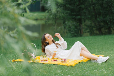 Beautiful young woman in a white dress drinking wine in the garden on a picnic