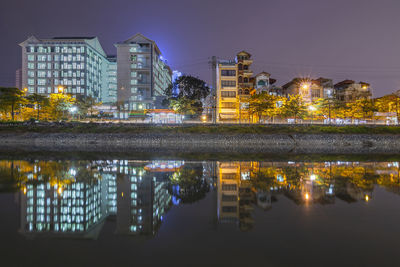 Illuminated buildings by lake against sky in city at night