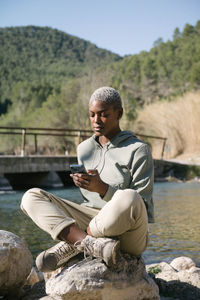 Full body of african american female sitting with crossed legs on rock and browsing cellphone on coast of rive flowing in hilly terrain