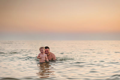 Portrait of couple swimming in sea against sky during sunset