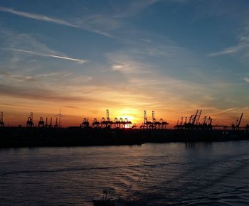 Silhouette cranes at harbor against sky during sunset